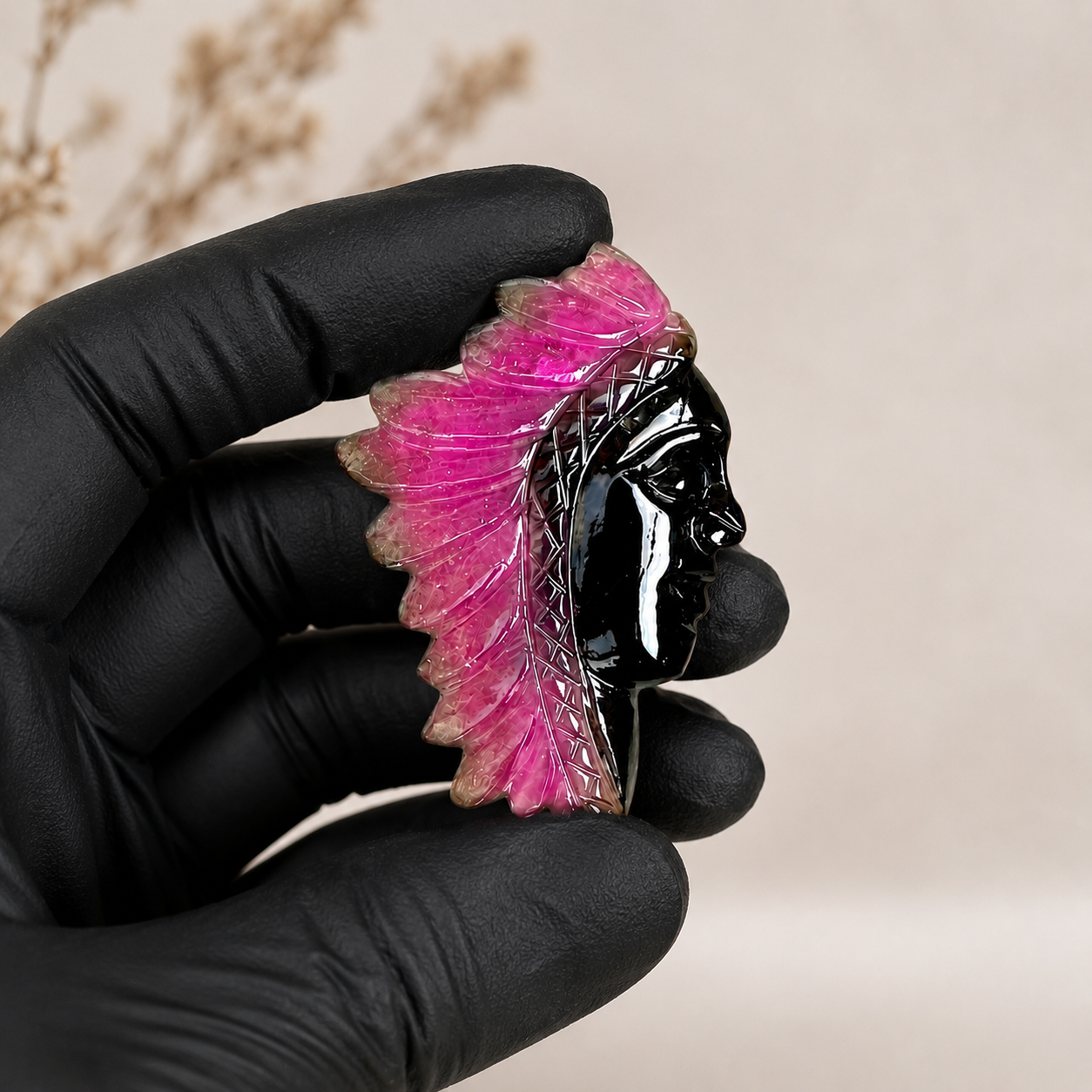 Black gloved hand holding a pink and black handcrafted gemstone chief head carving with detailed feather design.