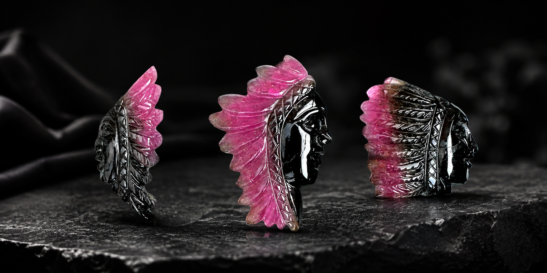 Three handcrafted pink and black gemstone tribal face carvings displayed on a dark textured surface with detailed feather-style crystal artwork.