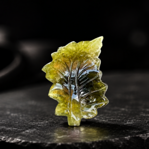 Hand-carved translucent green gemstone leaf placed on a dark stone surface with a luxury black background.
