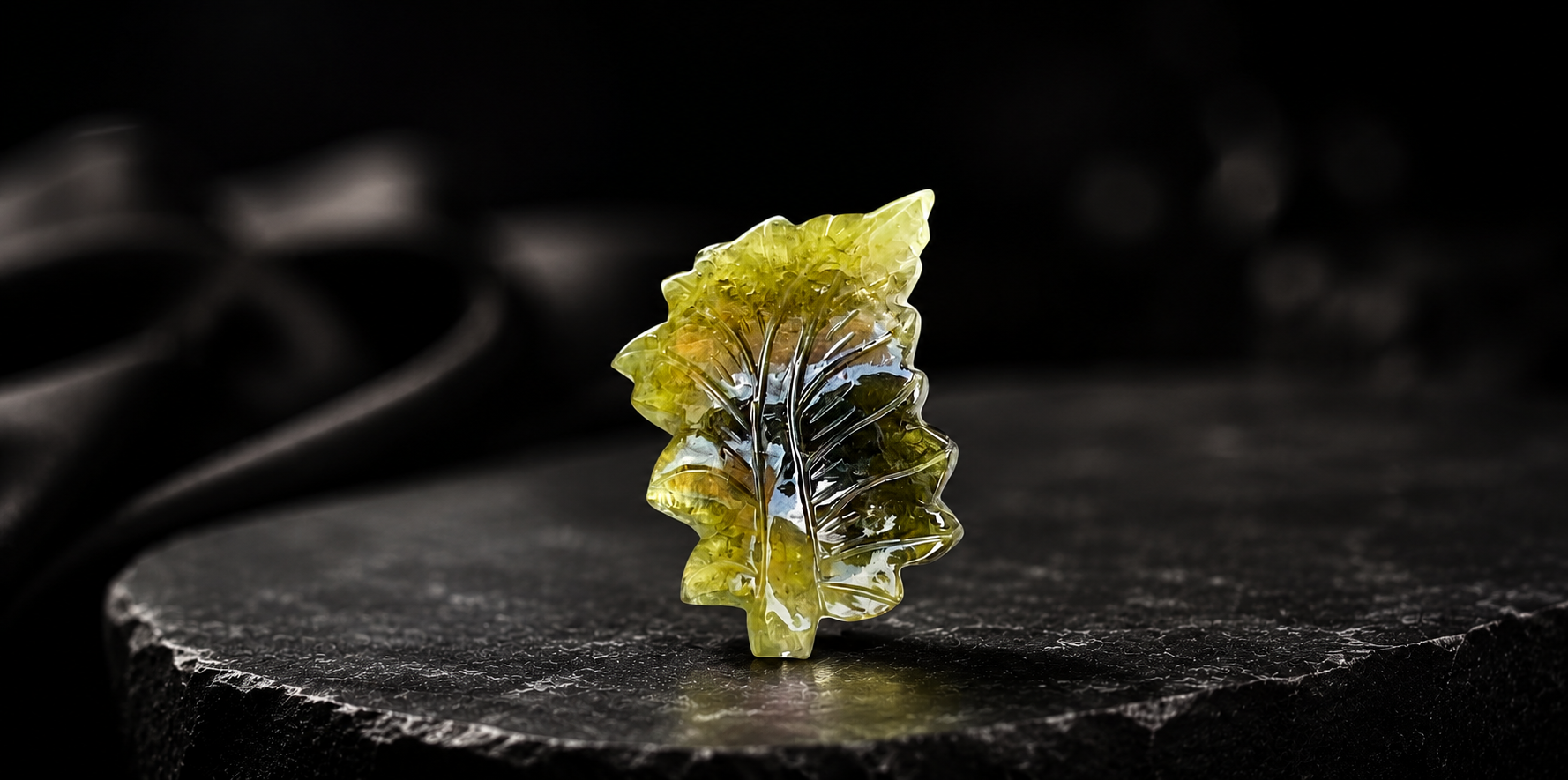 Hand-carved translucent green gemstone leaf placed on a dark stone surface with a luxury black background.